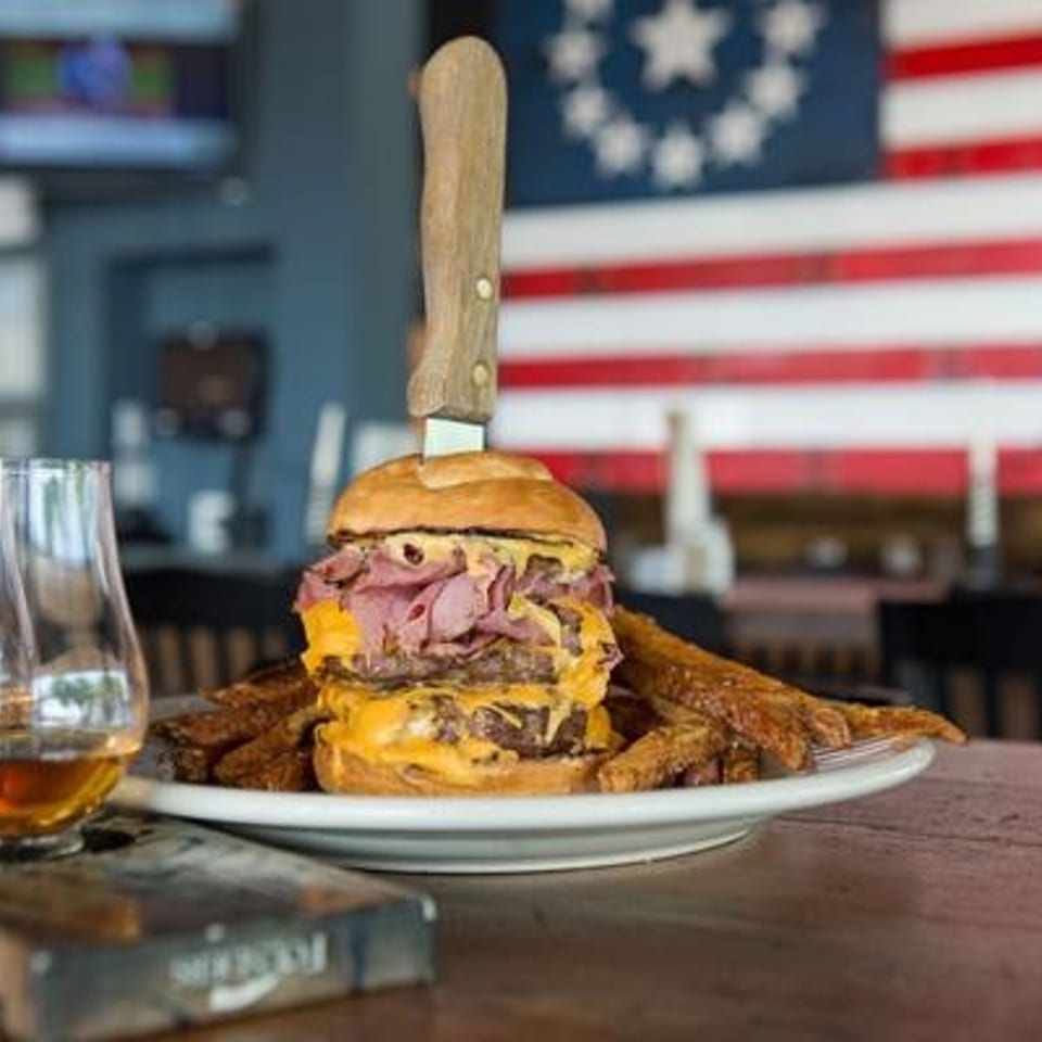 Stacked cheeseburger with pulled pork and fries on a plate, alongside whiskey glasses, in a restaurant with an American flag.