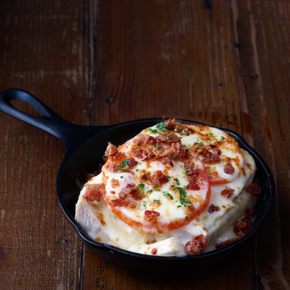 Cheesy baked dish topped with tomato slices, crispy bacon, and herbs in a black skillet on a rustic wooden table.