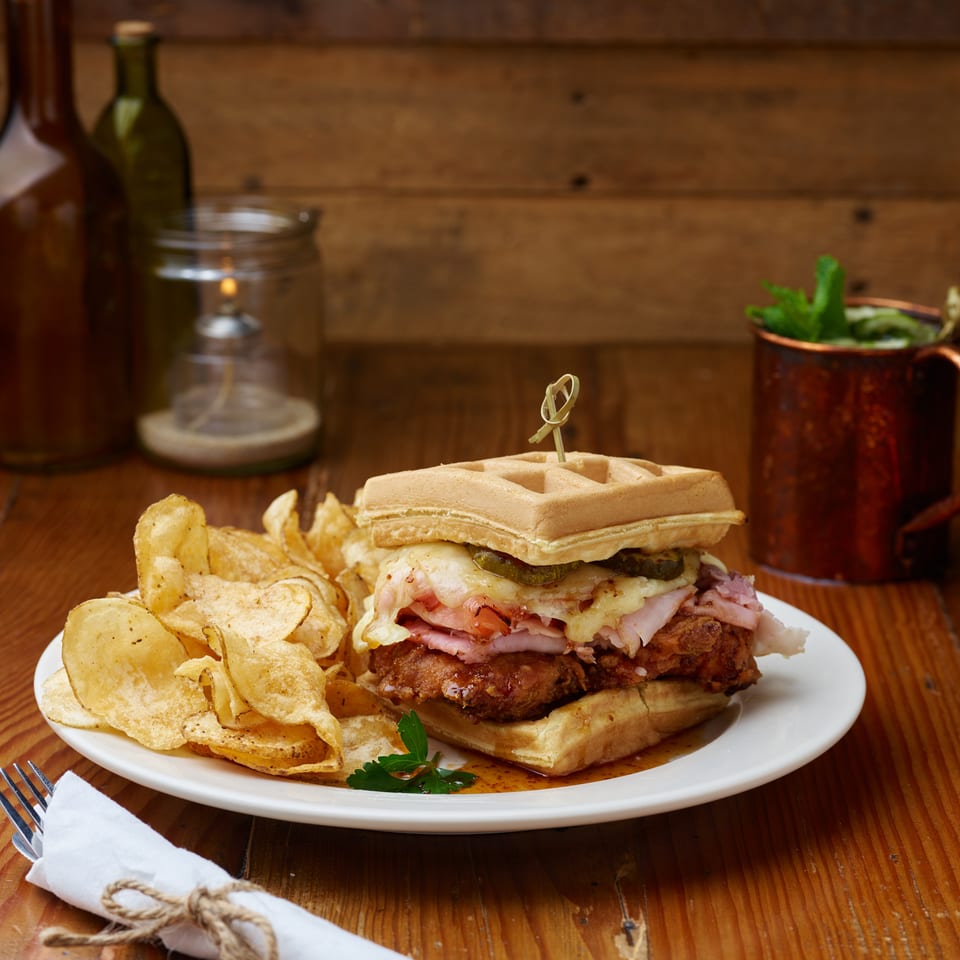 Waffle-topped fried chicken sandwich with melted cheese and ham, served with potato chips on a white plate.