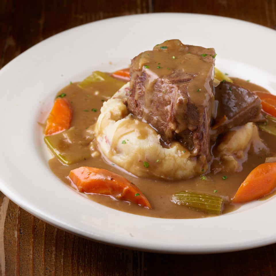 Braised beef topped with gravy, mashed potatoes, and carrots and celery on a white plate.
