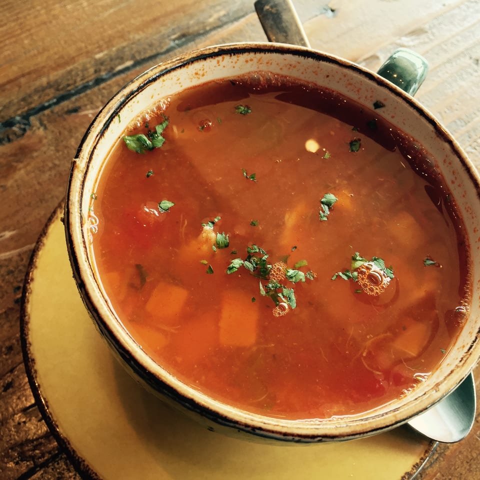 Bowl of reddish vegetable soup garnished with fresh herbs, served on a saucer beside a spoon atop a rustic wooden table.