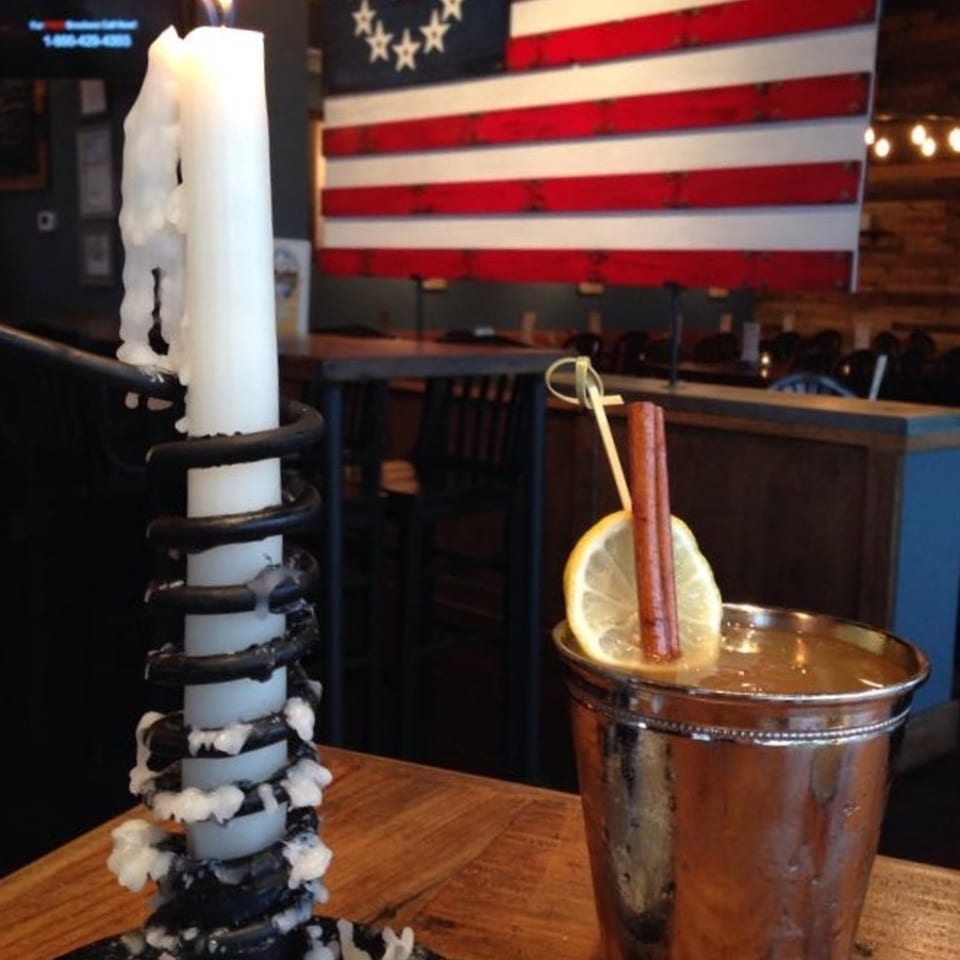 A melting candle and copper cocktail garnished with lemon sit on a wooden table beneath an American flag mural in a restaurant.